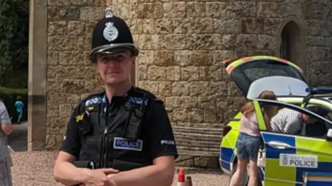 West Mercia Police A man in a black police uniform with a black police helmet in front of a large stone building and a police car which two people are looking inside.