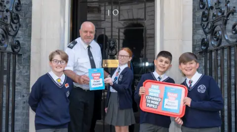 Four 11-year-old schoolchildren, three boys and a girl in blue uniforms, hand a card to a police officer on the steps of Number 10