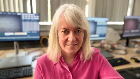 A long-blonde haired woman wears a pink shirt with an open collar. Behind her is a brown desk with three screens on it. She had brown eyes and smiles to the camera. The white blinds behind her are shut, they sit above the desks. 