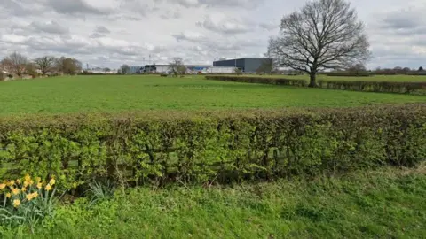 Google Pictured is a green field behind a hedgerow with some industrial-looking buildings at the back