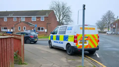 A blue and yellow marked police crime scene investigation van parked on the side of a road beside a row of terraced red brick houses. A black car is parked in front of it. The sky is grey.