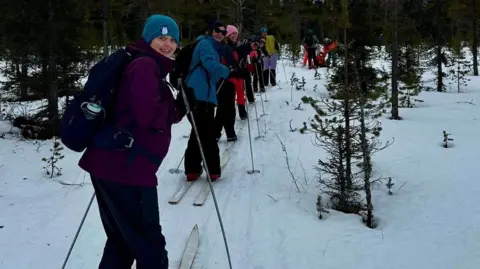 University Hospitals Coventry & Warwickshire NHS Trust People in a line with ski equipment look back to face the camera. They are smiling, wearing hats and have bags on their backs.