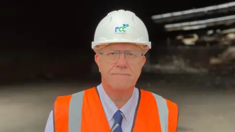 SAM READ/BBC A head and shoulders photo of Brian Ashby. He is looking at the camera with a slight smile. He is wearing an orange high-visibility vest and white hard hat which says FCC on it. The background is blurred but he is standing in front of a burnt-out industrial unit.