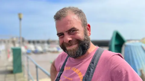 Amanda White/BBC Fisherman Peter Sanderson stands near the boats at Bridlinglton harbour