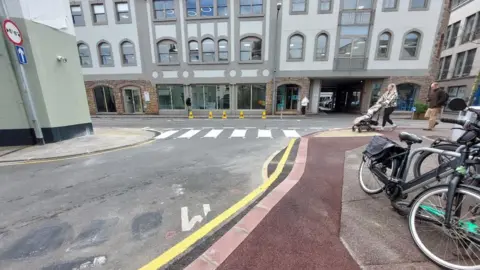 A mock-up of a zebra crossing at the end of a town centre junction. It is surrounded by quite high buildings, and there are pedestrians and bicycles nearby. 