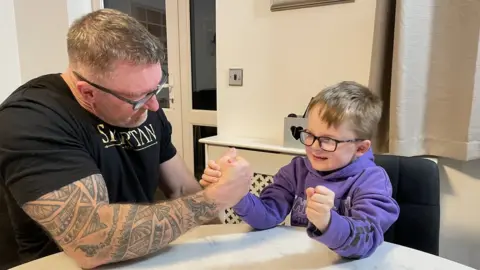 Sam Read/BBC Mark Waldon arm wrestles his 7 year old grandson Daniel on a dining table