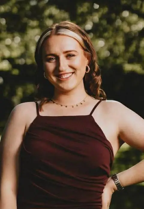 Summer Mace A woman with ginger hair and blond highlights smiles at the camera. She is wearing a burgundy dress, a necklace and hoop earrings. She is standing in a garden with green foliage behind her.