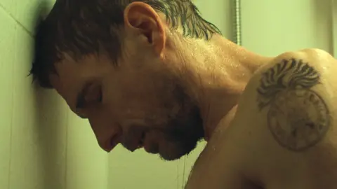 Bulldog Film Releasing Craig, who has short brown hair, sweating profusely and leaning his head against a white tiled wall. He has a tattoo of some sort of emblem on his arm.