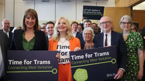 Tracy Brabin and Rachel Reeves stand in front of a crowd holding placards which read "Time for Trams" and "Let's get spades in the ground by 2028".