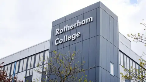 Against a cloudy sky, there's a tall grey block building. Windows run along two sides. At the corner between the two sides, a taller dark grey block has the words Rotherham College on it, in large white letters. There are trees in leaf at the far left and right of the picture and in the middle, which reaches up to just below the name of the college