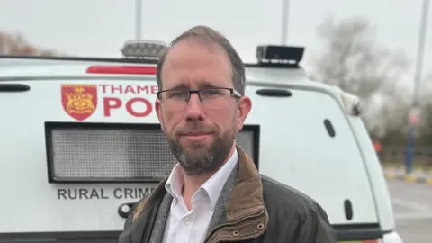 BBC Matthew Barber is standing in a green, waxed jacket in front of a white police van.