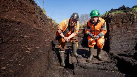 Two male archaeologists working at an excavation site. They are both kneeling down and wearing orange high vis suits, wellies and hard hats.