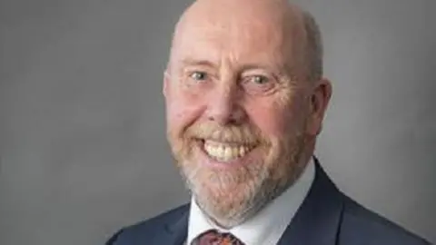 Cheshire East Council Headshot of Reg Kain looking at the camera and smiling. He is wearing a suit and tie. 