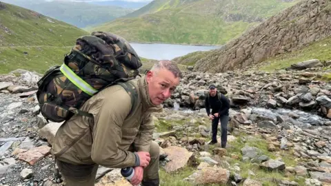 Scott Caswell Scott Caswell leaning against a rock on a hike with a military backpack on and florescent band around it. He is wearing a green jacket and trousers. Behind him a friend stands all in black with a military backpack on too.