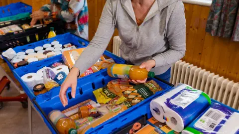 A medium shot of a food bank being run by volunteers at a community centre. Food that has been donated is lined up in crates on the table.