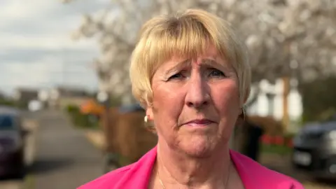An older woman with short cropped blonde hair and a bright pink jacket standing in a residential street of 1960s houses. She is looking at the camera and the background is blurred.