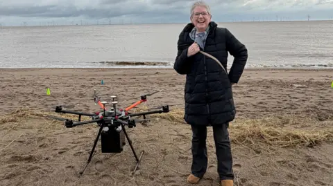 Jenny Fox stood on a sandy beach with the sea behind her. She is wearing black jeans, brown boots, a long black padded coat and a colourful scarf. She has short blonde hair and is wearing glasses. She is smiling at the camera. On her right is a large drone containing her husband's ashes. The sky is overcast.