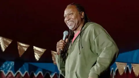 Getty Images Reginald D Hunter on a stage wearing a green hoody holding a microphone