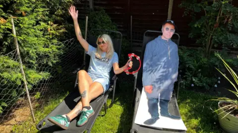 Liv Bridge reclines on a garden chair while holding a tambourine in her arm. Next to her is a cardboard cut out of Liam Gallagher. Liv is blonde, and is wearing a Oasis band t-shirt and dark sunglasses.