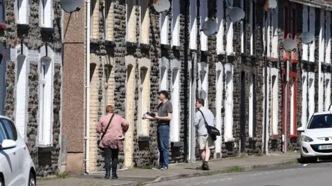 Getty Images Three people canvass knock doors in a terraced street holding leaflets.