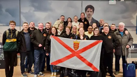 A band in an airport,  grouped together, with the people in the front holding a Jersey flag