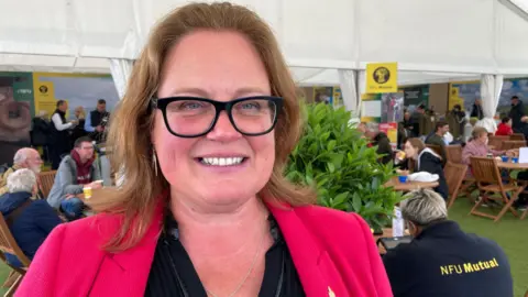 Farmer, and NFU Vice President, Rachel Hallos. She is standing in a marquee at an event. She is wearing a pink blazer and smiling. 