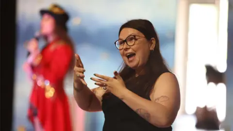 A woman acting as an interpreter, with her hands moving as her mouth is open. She has brown hair and wears a black shirt, with tattoos on her arms. In the background, a woman in a red coat holding a microphone is blurred.
