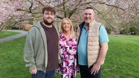 North Yorkshire Council The image shows three people standing together in a park, smiling at the camera. They are positioned beneath pink blossom trees, with green grass and winding paths visible behind them. The man on the left is wearing glasses, a dark t-shirt, shorts, and a green hooded jacket. The woman in the middle is wearing a pink and purple patterned dress, while the man on the right is dressed in a light blue t‑shirt with a beige padded gilet.
