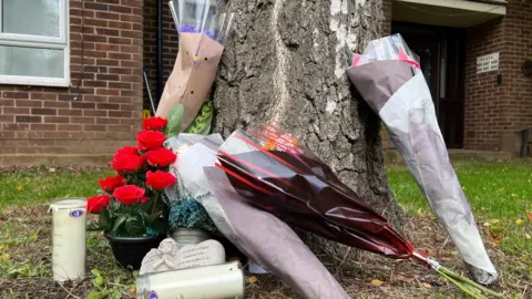 Stuart Woodward/BBC Floral tributes, candles and cards left for Kieran Shepherd resting on and next to a tree on a grassy patch in front of housing