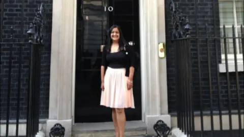 Sanju Pal Sanju Pal, wearing a black top and a light coloured skirt, standing on the steps of the Prime Minister's residence in Downing Street.