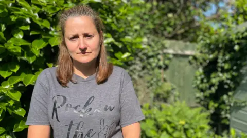 Emily Fryer, with tied-back brown hair and a dark-blue t-shirt, stands in front of a green bush in her garden and is looking solemnly at the camera