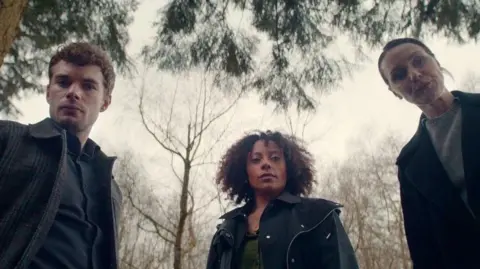 Netflix Catherine Ayers looks down at the camera along with two other characters in Missing You. They appear to be standing in a wood of forest as you can see trees behind them.