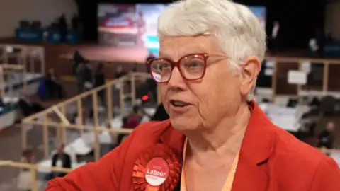 An older woman with short white hair, wearing a red blazer, red glasses and a red rosette with 'vote Labour' written across it stands against a railing.
