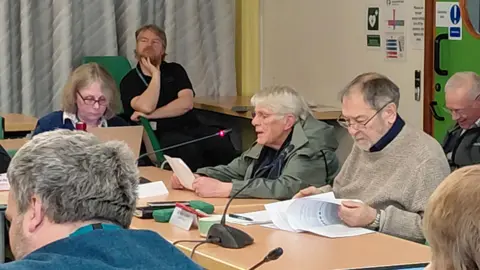 LDRS An elderly woman in a green anorak reads from some notes at a meeting. A number of people sit nearby looking at reports or checking laptop screens. 