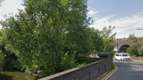 Google White campervan about to cross the River Rother at Catcliffe on the B6067 Treeton Lane. Double yellow lines along the brown brick bridge and the River Rother with trees and bushes  to the left. Blue sky and white clouds above, and shadows from riverbank trees on the road