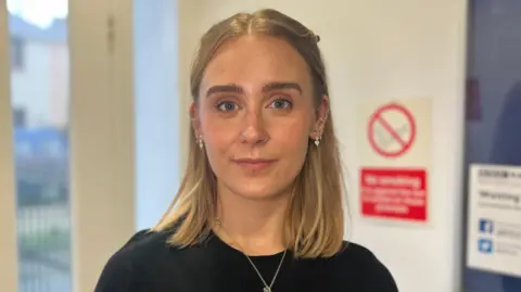 BBC Aimee is a young woman with shoulder length blonde hair. She is wearing a black t-shirt, a silver necklace and silver earrings and is looking into the camera. She is stood in front of a door and white wall with a no-smoking sign on it out of focus behind her.