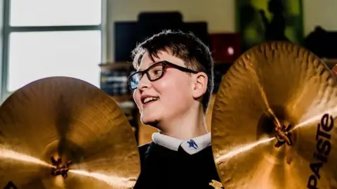 Antonio Piazza A young boy with short brunette hair stands between two symbols. He is wearing thick, dark rimmed glasses and has a white collared shirt on underneath a dark jumper.