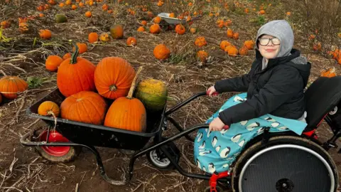 A boy sits in a wheelchair while holding the handles of a wheelbarrow filled with pumpkins. He smiles at the camera, wearing a black coat and a grey hoodie with the hood up, with a blue blanket with penguins on it wrapped around his lap. He is in a field filled with pumpkins.