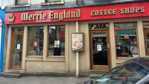 The exterior of a building with a red sign in gold writing which reads Merrie England coffee shops'.