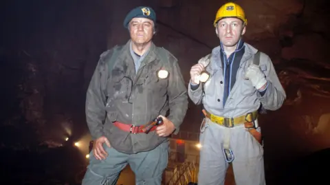 The actors Joe Don Baker and Bob Peck are dressed (in character) in caving clothing and equipment and are standing in a cave which is artificially lit by huge lamps. A portable cabin can be seen in the background.