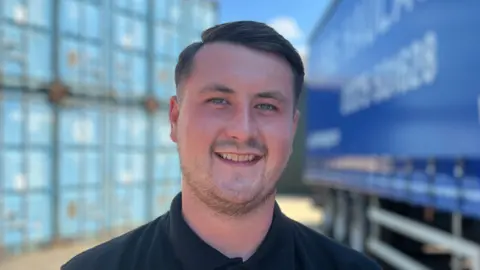 Simon Dedman/BBC Frankie Stonebrook has short dark hair and is smiling while wearing a black polo shirt. He is standing in front of large blue containers and a blue lorry.