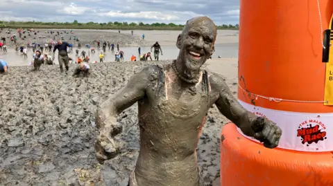 Lewis Adams/BBC Man covered from head-to-toe in mud. Lots of people behind him can be seen making their way across a very muddy river estuary.
