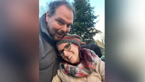 A man with dark brown hair and dark facial hair is wearing a grey coat, and has his arm around a woman. She has red hair, a knitted hat, black glasses, a knitted scarf and a beige coat. They are posing in front of a christmas tree with lights on it
