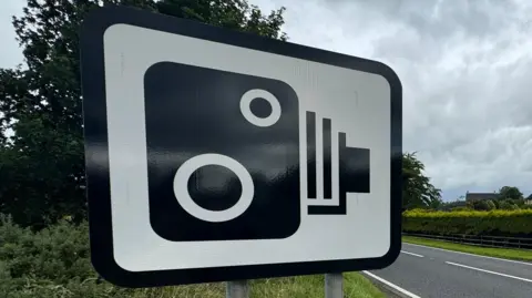 BBC A white road sign with a black speed camera symbol. It has a black border. Behind on the right is a road with green hedges above. To the left is green grass and trees.