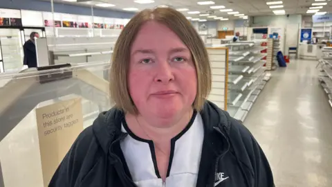 Sheelagh Gray - a woman with chin-length blonde hair looks at the camera. She is standing in the middle of a Boots store, with empty shelves all around her. She is wearing a black, Nike jacket and a white Boots shirt.