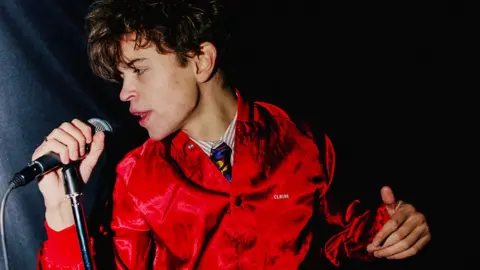 UMG A studio shot of a young man in a red, velvet jacket holding a microphone on a stand and leaning in as he sings a note.