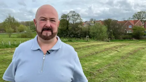 Man in light blue zip up polo shirt stands on grass in a garden. He is bald headed and has a beard.