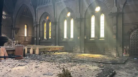 Image shows the fire damaged interior of a church. All over the floor is dirt and parts of the roof which fell in. There is black ash everywhere as well. The sunlight is still shining through the stain glass windows and down from the damaged roof. There are some light brown church pews which seem relatively undamaged on the front right hand side but to the left are some more pews which look more damaged. 