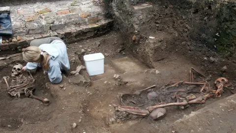 Skeletal remains lying in the soil bordered by a brick wall. A woman is on the ground next to one of the skeletons and there is a white bucket at her side. The woman has long blonde hair tied in a ponytail and is wearing a blue jumper and grey trousers. 