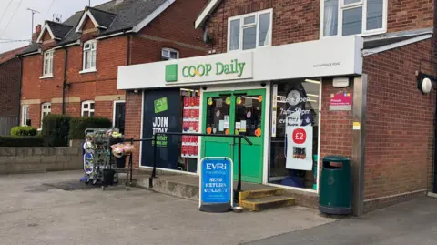 A small co-op daily store next to some homes. It has a white and green sign and signs for parcel delivery outside on the pavement. 